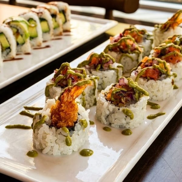 Two white platters of sushi rolls, one with avocado and the other with shrimp tempura and sauce.